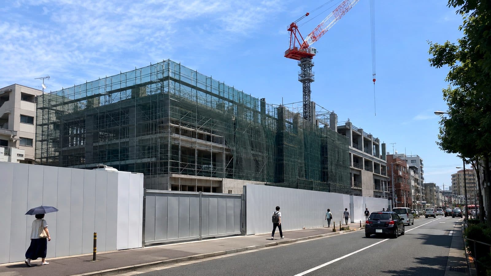 街路に沿って仮囲いが続き、足場とタワークレーンが立つ建築工事現場のイメージ